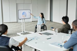 Group of colleagues at a table while a woman in a wheelchair points to a presentation on a TV monitor.