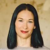 Headshot of Angie Barker. She has dark, shoulder length hair. The tan background is intentionally blurred.